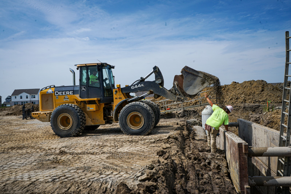 John Deere wheel loader moving soil beside a concrete drainage structure at a subdivision construction site, with a worker inspecting the structure.